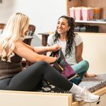 Two girls sit on a bed, brunette smiling, blonde going through backpack