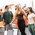 Blonde and brunette women stare hopefully upward as brunet man sings at their side