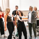 Curly-haired brunette woman holds out a large cake, blonde woman appreciates it as ensemble sing around them