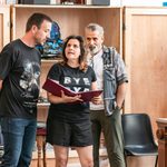 Nina Wadia performs, binder open in her hands, two men peering over her shoulders