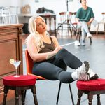 Blonde woman dreamily writes in notepad, recling in plastic chair, feet up in red stool, martini on another red stool