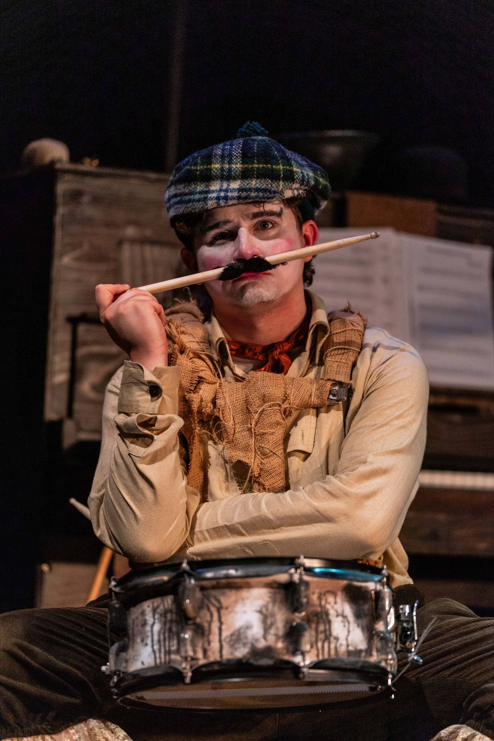A man with a drum holds a drumstick with a moustache attached to it to his face.