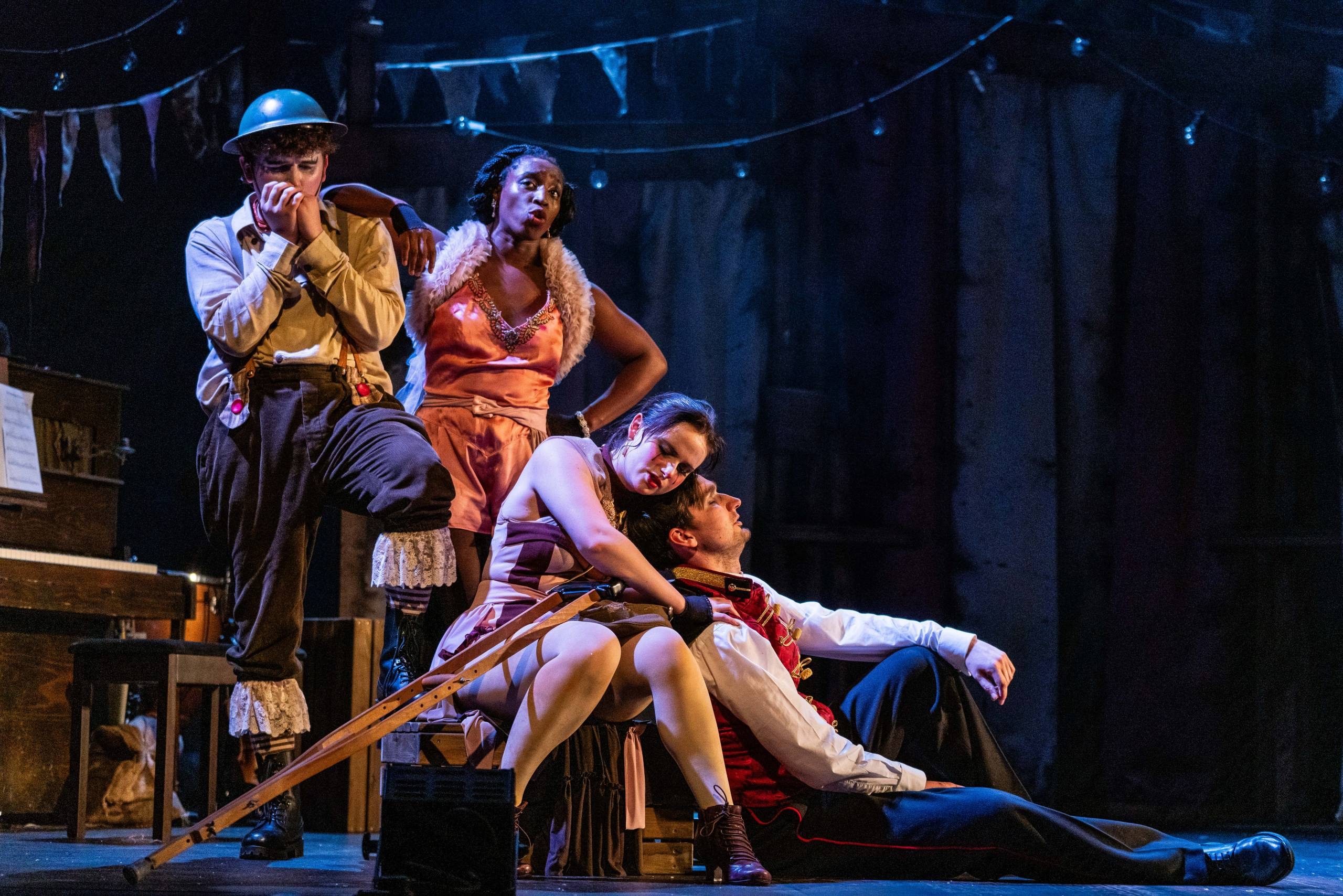 Four characters on stage, two male and two female. One male stands and plays the harmonica while one female has her arm on his shoulder. The other male and female sit on the floor together.