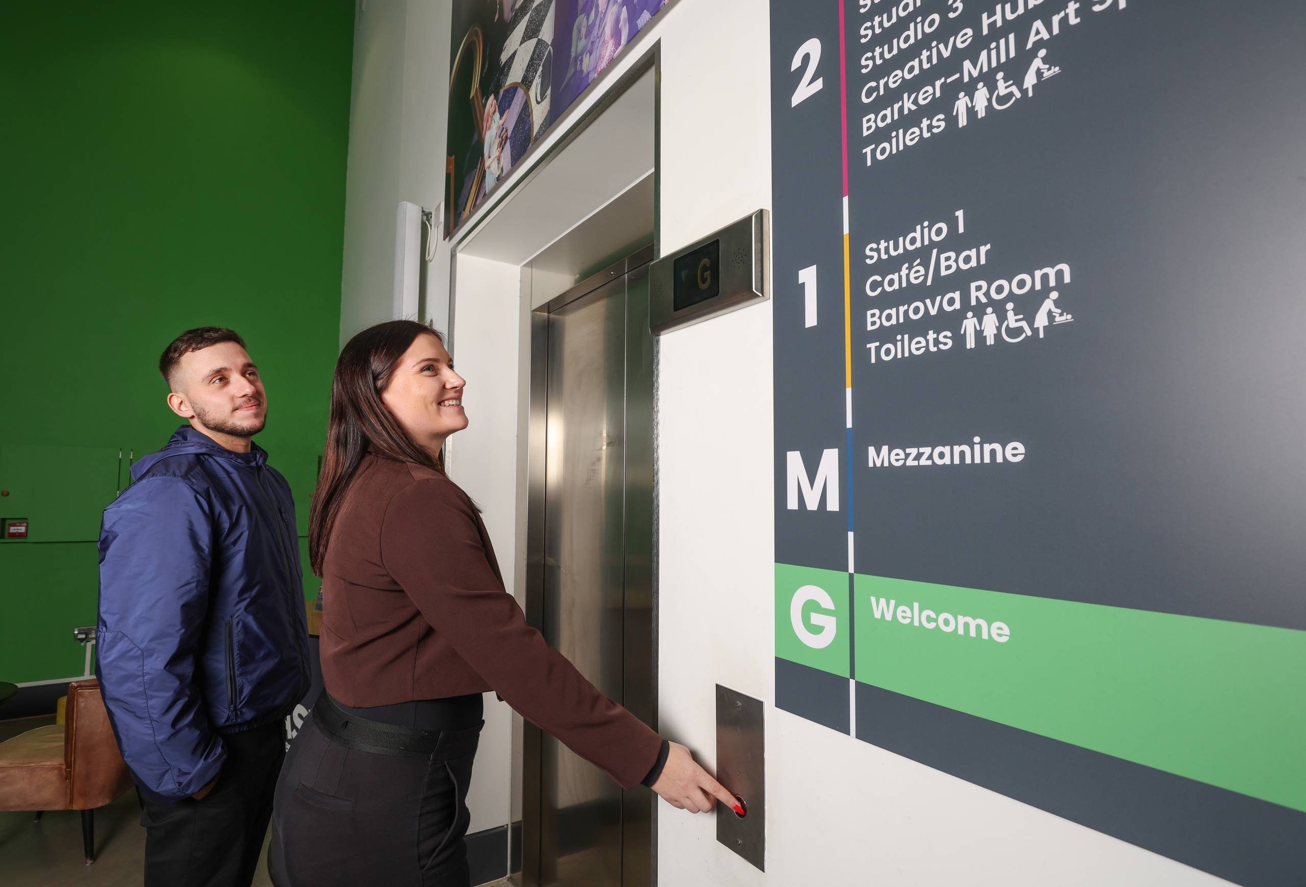 A woman stands in front of a lift pressing the button to call the lift. To her right is a sign showing all the areas in the venue. To her left is a man looking up at the sign.