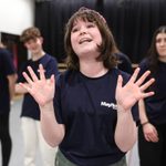 A young participant of our Youth Theatre is smiling and holding both hands in the air.