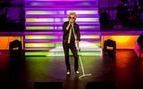 Rod Stewart impersonator wearing black with gold tie singing into standing mic on purple and green lit stage