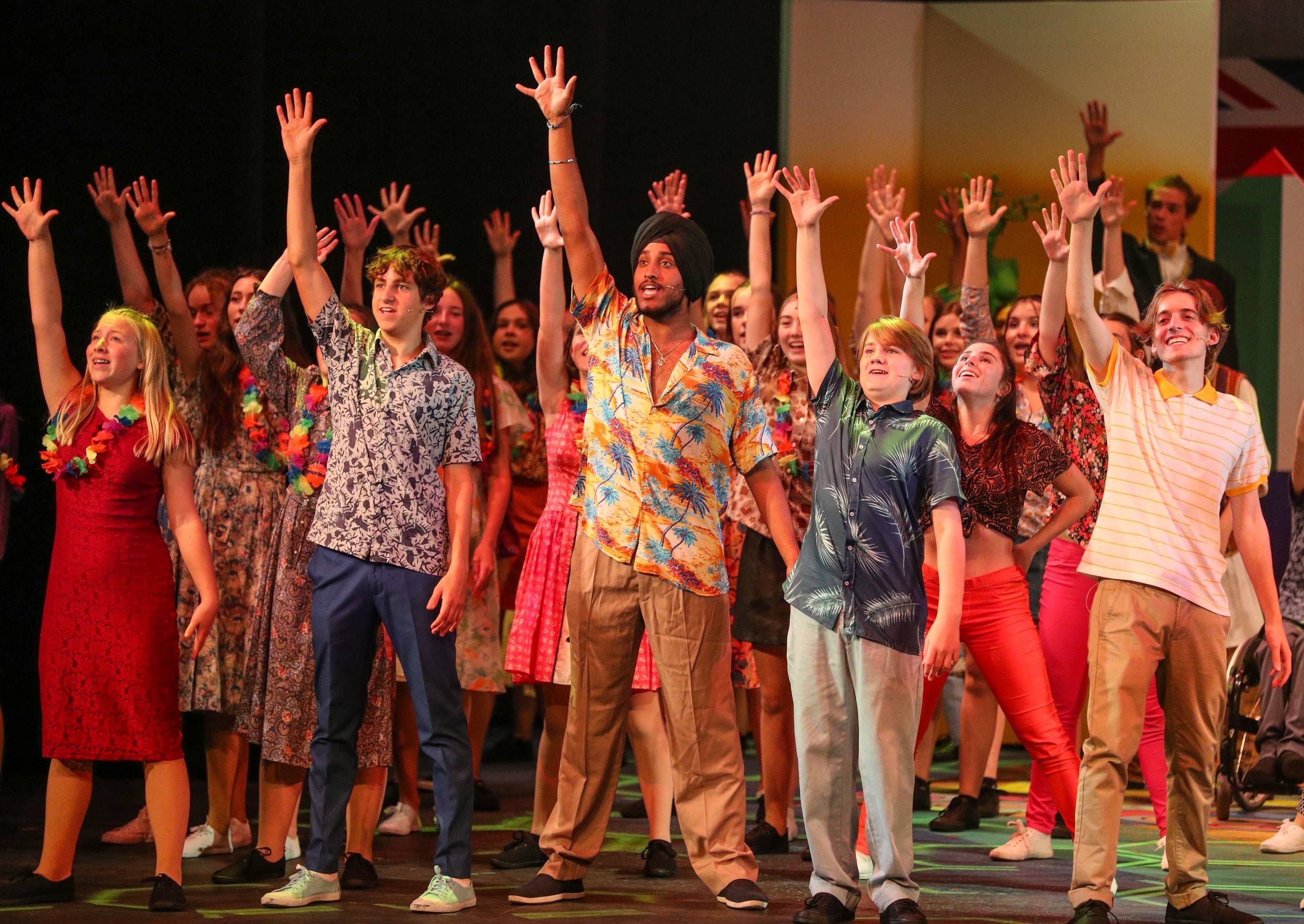 The cast of Summer Holiday raise their right arm in the air with their hands spread open. They are wearing summer shirts and dresses.