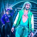 Triton (Thomas Lowe) in green leotard dipping down to his tulle tutu with green letterman over the top, stares outward. Behind him, Ursula (Shawna Hamic) in purple jean jacket, glasses and purple hair, looks at him.