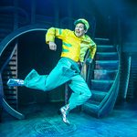 Eric leaping with a smile, wearing silver , heeled boots, teal trousers, and yellow jacket and hat.