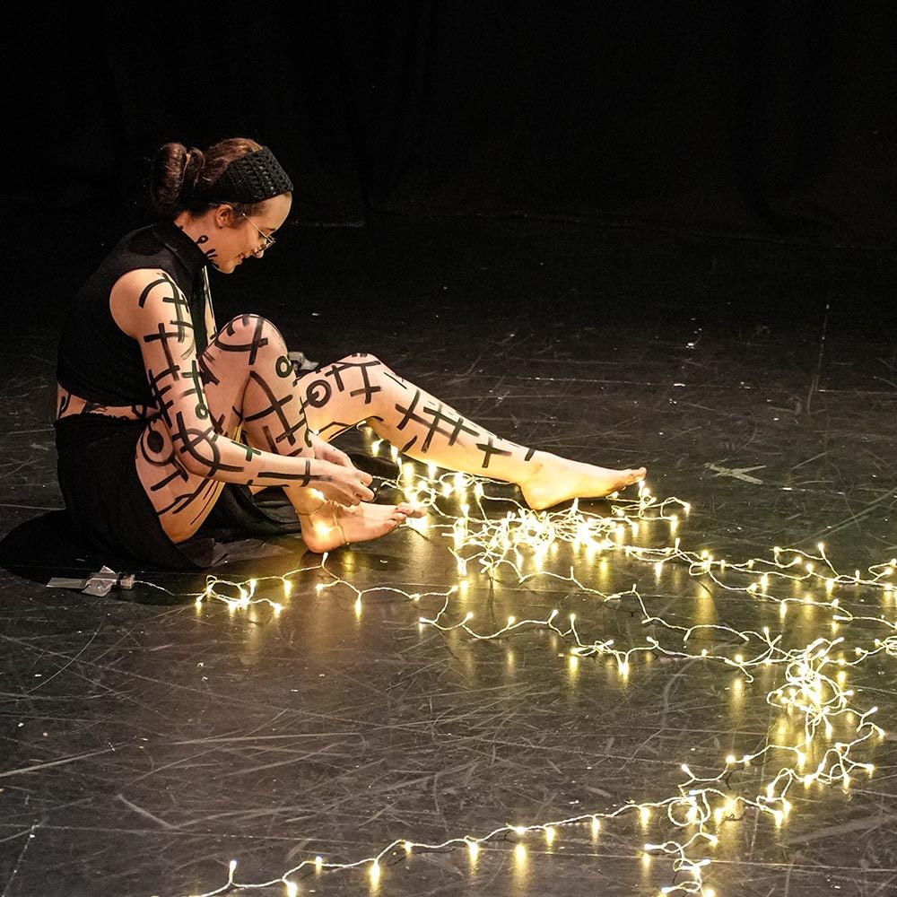 Young white female performer sat on stage surrounded by a string of fairy lights