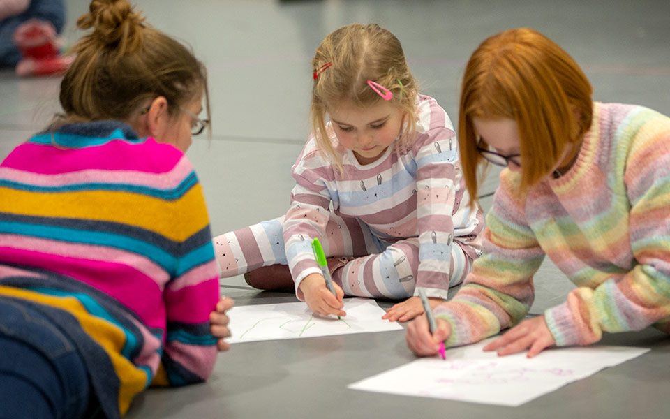 Lying on the floor, a mother in multicoloured striped jumper watches her two young daughters in colourful jumpers drawing on sheets of paper.