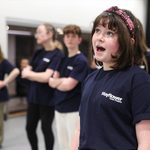 A young girl wearing a Mayflower Youth Theatre branded t-shirt and pink headband sings in front of peers