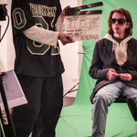 Young boy in front of someone holding a clapper board. The boy is wearing sunglasses and a coat and is sat on a chair in front of a green screen.