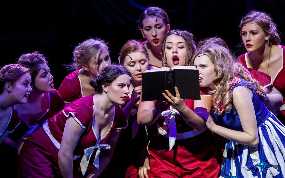 Young female actors singing on stage whilst looking at a book from our 2017 Summer Youth Project Guys and Dolls.