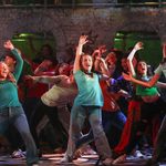 3 female dancers taking centre stage surrounded by more dancers. All 3 female dancers have their hand or hands raised and are wearing t-shirts and jeans.