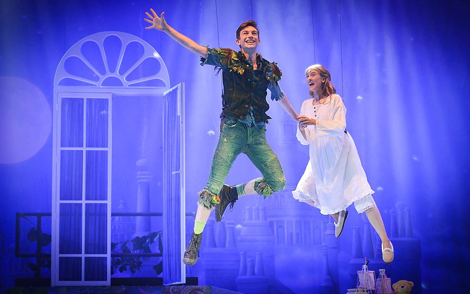 Young actors flying in a scene from Peter Pan - An Awfully Big Musical Adventure. The boy is wearing dirty green denim shirt and top and the young girl is in a white night gown.