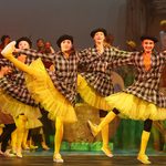 A group of young white dancers wearing yellow leggings and black and white checked coats with their right leg raised and their arms strecthed.