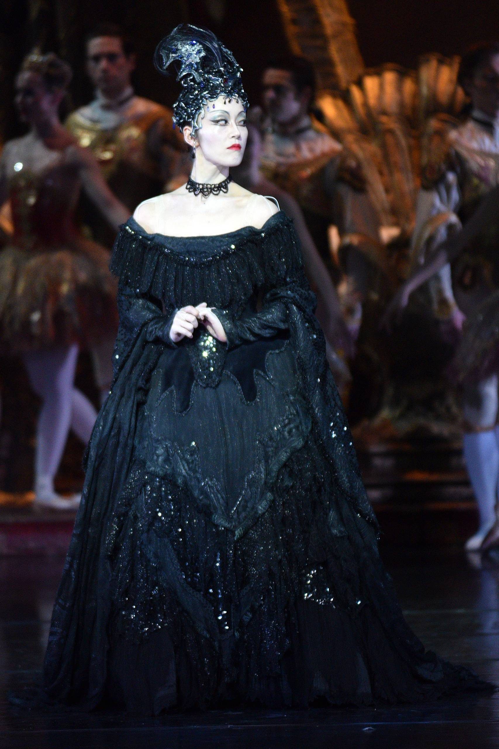 The evil queen stands tall and wears a long black dress. Ensemble dancers are in the background wearing white and gold costumes.