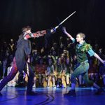 Two young men in costumes engaging in sword fighting on stage during a performance of Peter Pan - An Awfully Big Musical Adventure.