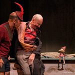 Two men wearing a mixture of red, grey and brown tattered clothing look down at a Pinocchio puppet