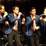 Four men in blue blazers and red ties leaning together to sing into hand mics