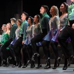 Riverdancers in green and blue stand in a line, hand in hand, Irish dancing.