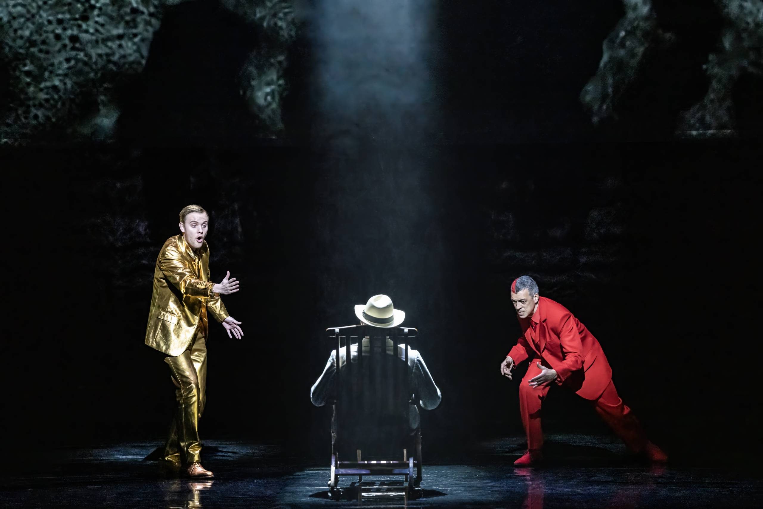 Apollo (Alexander Chance) in gold suit and Dionysus (Roderick Williams) in red suit, sing at Ashenbach facing away in wooden chair.