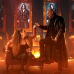 Vampire Steve Steinman holds his hand out to a white long-haired brunette sitting on a chaise lounge in candle and fire lit room overlooked by four large paintings of vampires.
