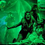 Vampire woman bares her fangs over a terrified woman in a wedding dress on the floor in her arms, the scene washed in green light.