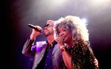 George Michael Impersonator side hugs a black woman with curly grey hair, both singing into hand mics
