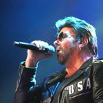 Close up side view of George Michael Impersonator singing into hand mic wear leather jacker with BSA on the breast zip up pocket