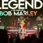 Bob Marley performer in green, yellow, red jacket and hat throwing hand into air on stage