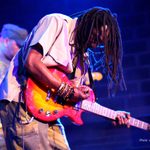 Bob Marley performer in plain shirt, dreadlocks free, bent over the guitar he's playing