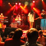 Bob Marley performer in plain shirt and dreadlocks hanging free walking across stage with band towards female singer in blue skirt and headscarf