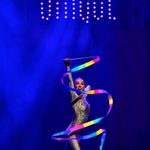 Woman in silver leotard waving rainbow ribbon in front of her on dark stage with CIRQUE in lights up high.