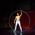 Bare-chested white brunet in white trousers stands in the middles of a large hoop, holding it up with one hand.