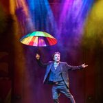 Mime with suit over white and black striped top holds open a rainbow umbrella, gesturing as though feeling for rain in the multi-coloured spotlights.