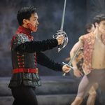 Tybalt (George Liang) holding rapier and dagger, face covered in blood, wearing black and red tunic