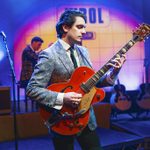 A man in a patterned suit is playing a vintage red guitar.