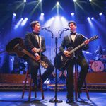 Two men are sat on stalls wearing tuxedos and playing guitars while they sing into microphones.