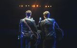 Two men wearing sparkly black suits have their backs turned to the camera while they play guitars.