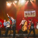 A man is singing into a old style microphone while he points into the air. Around him is a group of other men playing guitars. Behind him is a double bass player and a drummer.