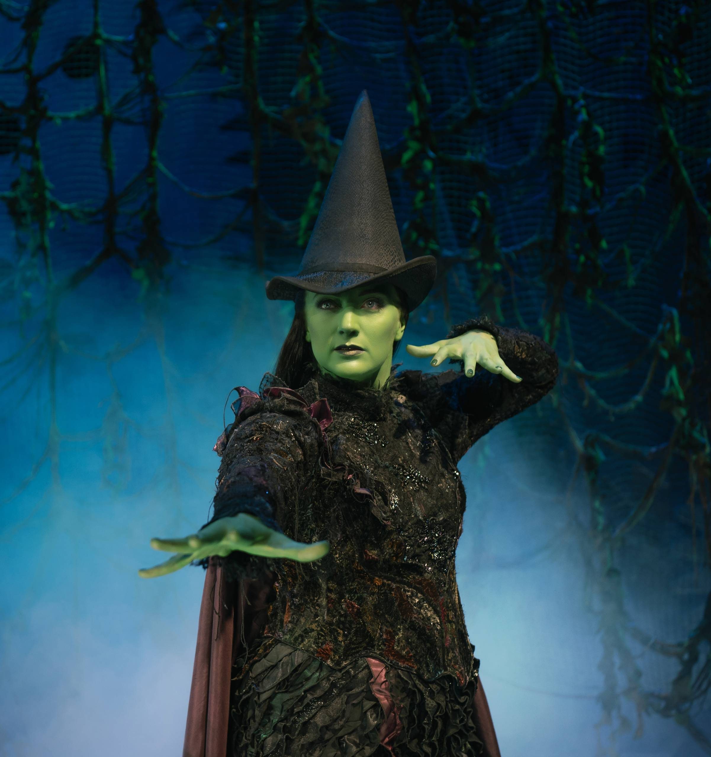 Laura Pick as Elphaba waving her hands as though casting a spell in a forest at night