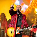 Steve Steinman in red leopard print jacket, holding wood-effect electric guitar, pointing out past the microphone. Two female backing singers behind.