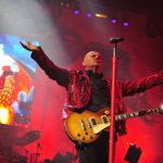Steve Steinman in red leopard print jacket, holding wood-effect electric guitar, hands wide mid-clap