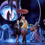 Lucy and Tumnus walk arm in arm below an umbrella. He holds a wicker basket. Figures in white dance around them and in a glowing circle in the backdrop a saxaphone player is shadowed.