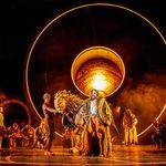 Aslan, a man in a fur coat and his life-sized lion puppet avatar, stand in the middle of an orange-washed stage as characters and musicians bow around him, 3 Pevensie children watching