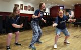 Mayflower Blue Apple Inclusive Youth Theatre Three girls in blue t-shirts and casual clothes practice dramatic poses in a room with wooden floors and chairs.
