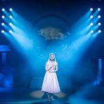 Sandy (Hope Dawe) in pale pink tea dress clutches her chest as she sings in spotlight