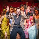 Vince Fontaine (Joe Gash) in leopard print jacket throws his hands up, ready to party, as the students gather excitedly around